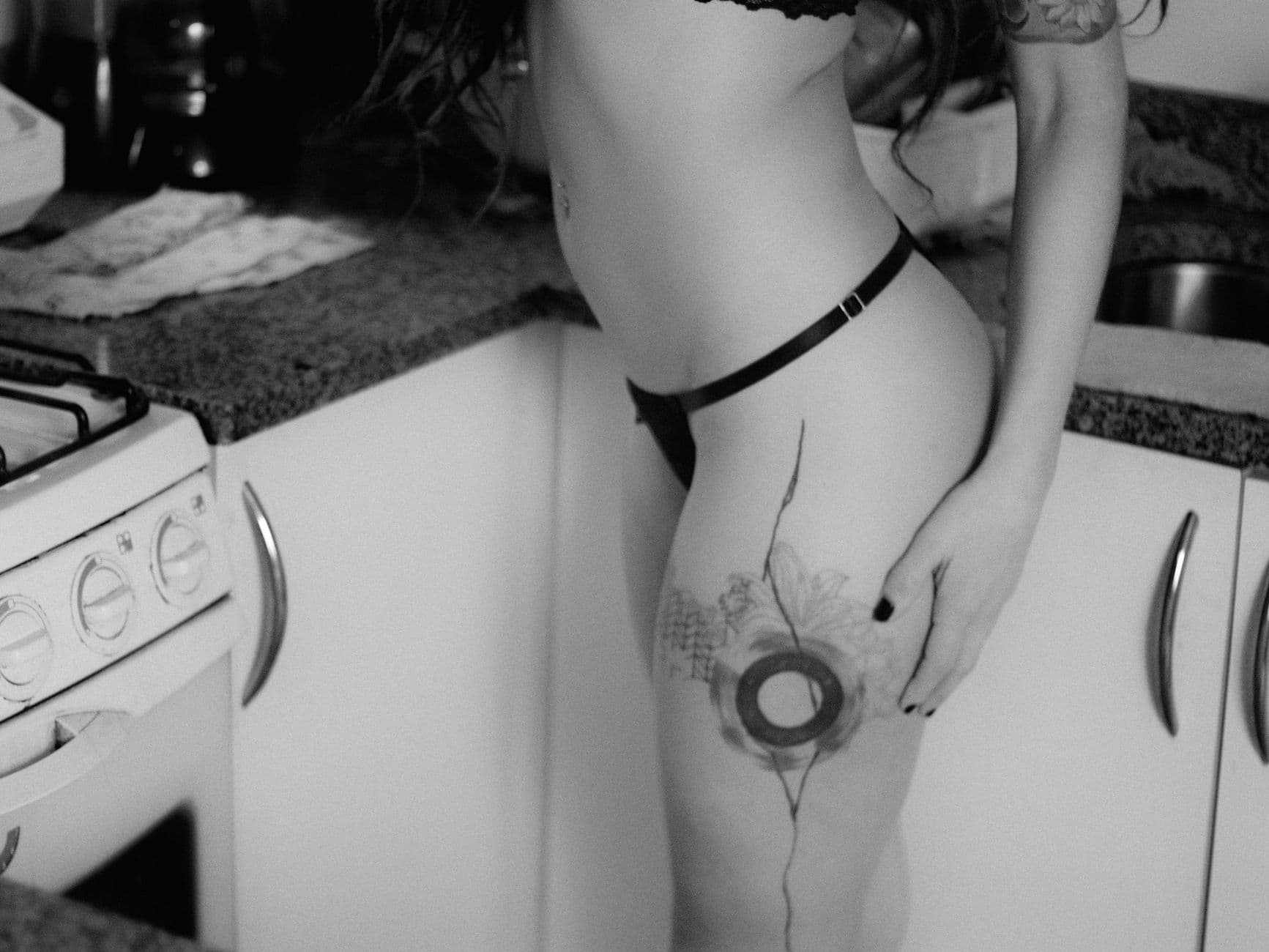 Black and white photo of a woman with thigh tattoo in a kitchen setting.